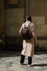 A beautiful fair-skinned model with long, dark hair is photographed from the back, showcasing a sophisticated brown leather backpack and a timeless beige trench coat.