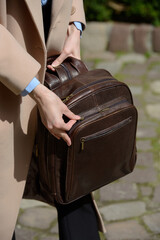 A stylish female model showcasing a brown leather backpack with detailed zippers, held against a sophisticated beige trench coat in an urban setting