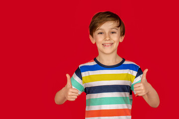 Portrait of a smiling little boy showing thumbs up isolated over red background. Little small smiling happy boy showing thumb up like gesture. Portrait of beautiful little boy giving you thumbs up