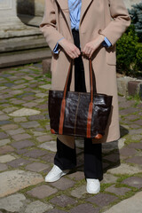 A stylish urban shot captures a woman in a beige coat and sneakers, showcasing a spacious leather tote bag with contrasting handles