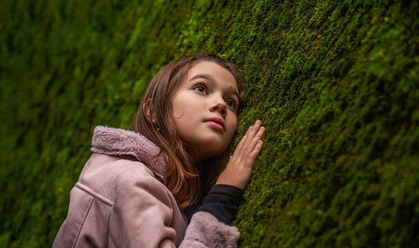 Cute little girl standing in deep dark forest at old tree covered with green moss. Girl is touching fluffy moss in summer forest. Child girl lies on the soft and delicate moss covering forest
