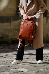A stylish female model showcasing a brown leather backpack with detailed zippers, held against a sophisticated beige trench coat in an urban setting