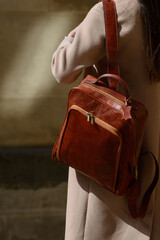 A beautiful fair-skinned model with long, dark hair is photographed from the back, showcasing a sophisticated brown leather backpack and a timeless beige trench coat.