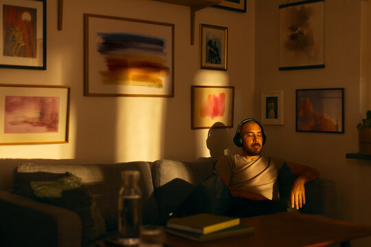 Man relaxing on a sofa with headphones in warm evening light. The cozy living room is decorated with framed artwork on the walls.