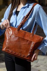 A professional close-up shot of a model showcasing a refined brown leather tote bag with unique braided handles and a decorative front pocket