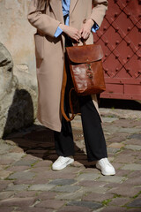 A stylish female model in a classic beige coat and sneakers showcases a sophisticated brown leather backpack with a unique flap closure and buckle detail