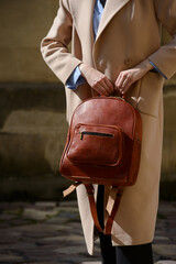 A stylish female model showcasing a brown leather backpack with detailed zippers, held against a sophisticated beige trench coat in an urban setting