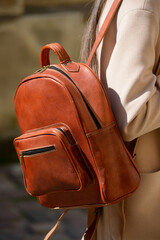 A beautiful fair-skinned model with long, dark hair is photographed from the back, showcasing a sophisticated brown leather backpack and a timeless beige trench coat.