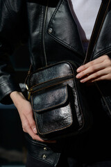 A stylish model in a black leather jacket showcases a unique black leather cross-body bag with a glossy sheen and a front flap pocket