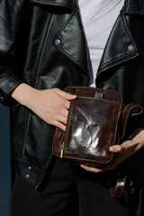 A stylish model in a black leather jacket showcases a unique black leather cross-body bag with a glossy sheen and a front flap pocket