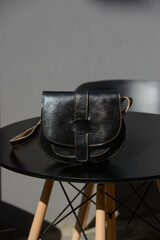 A small, black leather cross-body bag with a unique flap and contrasting stitching is showcased on a modern table in a sunlit outdoor setting