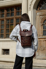 A stylish model is photographed from the back, showcasing a classic brown leather backpack and a trendy plaid overshirt against an urban backdrop