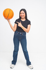 A confident young Asian woman in a full body shot standing and holding a basketball with one hand while pointing to it in an inviting manner. Isolated on a white background.