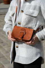 A stylish model in a plaid overshirt holds a small, reddish-brown leather bag with a unique flap and wide buckle, showcasing casual elegance