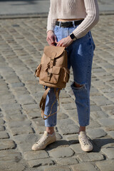 A stylish model in a cropped knit sweater and ripped jeans holds a refined leather backpack, showcasing a modern urban look