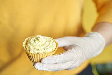 Female hand wearing glove holding yellow cupcake with swirled frosting against vibrant backdrop.