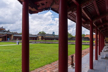 A colonnaded walkway with deep red columns frames a view of a vast green lawn and traditional...