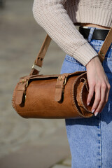 A close-up shot of a model in a knit sweater and jeans, showcasing a unique cylindrical leather bag with intricate side detailing and a belt.
