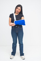An injured young Asian woman in a full body shot standing with an arm sling, grimacing in pain and discomfort. Isolated on a white background.