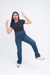 A surprised young Asian woman captured in a full body shot standing on one foot with arms in the air, mouth agape and eyes wide with astonishment. Isolated on a white background.