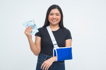 A pleased young Asian woman in a half body shot wearing an arm sling, smiling warmly while raising money with a content expression. Isolated on a white background.