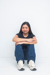 A relaxed young Asian woman sitting on the floor hugging her knees with a gentle smile, enjoying a chill and calm moment. Isolated on a white background.