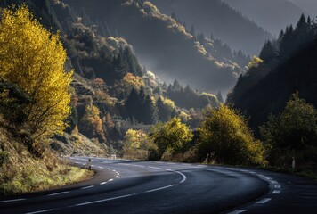 Winding road through a mountain pass in autumn, with trees showing vibrant golden and orange leaves against a misty mountain backdrop
