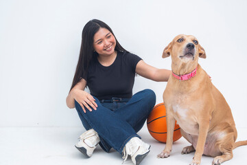 A playful young Asian woman sitting on the floor next to a basketball while petting a dog, smiling with joy and engagement. Isolated on a white background.