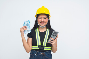 A cheerful young Asian woman captured in a half body shot wearing a safety helmet, holding her phone and money with a satisfied and joyful expression. Isolated on a white background.
