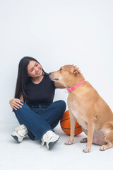 A cheerful young Asian woman captured sitting beside a basketball on the floor, smiling and gently petting a dog in a friendly manner. Isolated on a white background.
