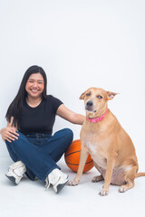 A lively young Asian woman captured sitting beside a basketball, smiling and laughing joyfully as she pets a dog on the floor. Isolated on a white background.