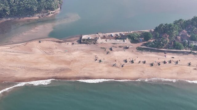 High-angle aerial view of a traditional fishing village on a sandbar between the ocean and a river at Kelus Kalvi Beach, India.