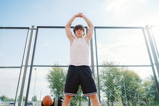 A young male basketball player stretches on a court under a clear sky. He prepares for practice, showcasing determination and focus. Nearby, a basketball rests on the ground.