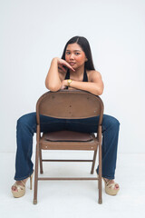 A posh young Asian woman sitting on a backward chair, posing elegantly with her hand gracefully resting on her chin. Isolated on a white background.