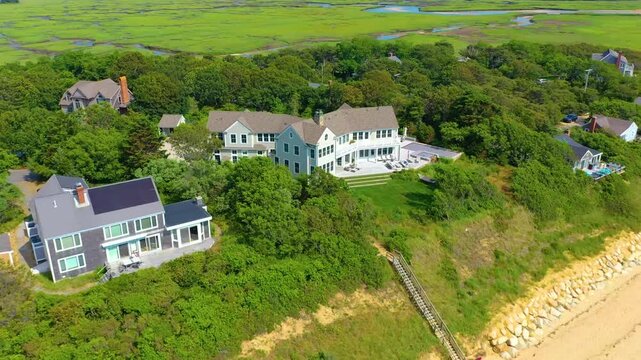 Scenic drone view of upscale houses along the shoreline, featuring manicured lawns, bluffside decks, sandy beach access points, and winding tidal marsh creeks flowing gracefully in the distance.