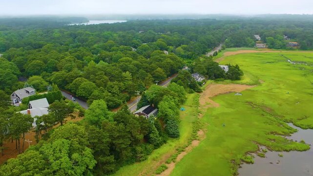 Aerial drone footage of Cape Cod marshlands, with winding tidal creeks flowing through lush green wetlands under an overcast summer sky, bordered by scattered homes and dense forest edges.