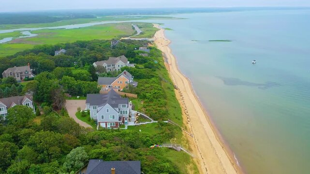 Cloudy day drone footage of a quiet coastal neighborhood along the shoreline, featuring sandy beaches, lush green marshes, winding tidal creeks, and beachfront homes set against the calm ocean.