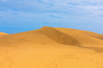 Desert dunes under blue sky. Wind has created ripples and sharp ridges on the sand surface. Footprints mark a path across the golden sand dune