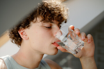 A young man with curly hair drinks a glass of water. Natural light filters in, creating a warm atmosphere. He seems refreshed and takes a moment to hydrate in a cozy space.