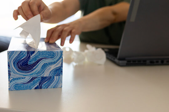 A sick person sits at a desk in front of a laptop computer. They are pulling a tissue from a box of tissues, likely to wipe their runny nose while working from home.