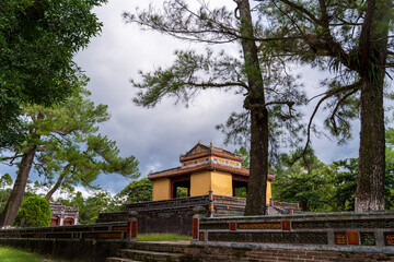A yellow and red pavilion stands on a brick platform and terraced steps, framed by tall trees under...