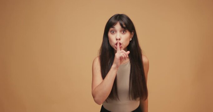 Beautiful hispanic woman holding finger on lips, blue studio backdrop. Pretty female with gesture of shhh, secret, silence, conspiracy, gossip, rumors concept