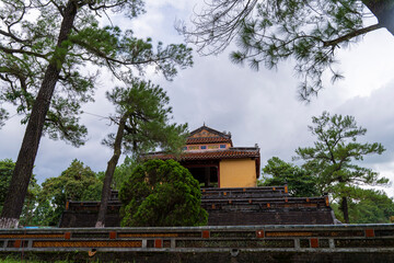 A yellow and red pavilion stands on a brick platform and terraced steps, framed by tall trees under...