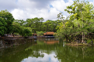 A tranquil lake reflects the Hien Khiem Pavilion and surrounding lush pine forest under a cloudy sky at Tu Duc tomb, Hue.