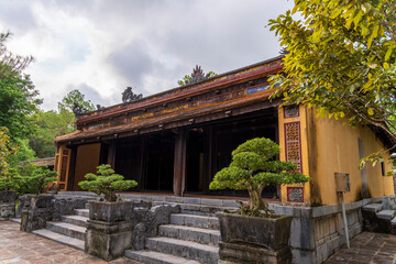 A traditional Vietnamese pavilion with yellow walls and a dark wooden interior stands on stone steps, flanked by potted bonsai trees under a bright sky.