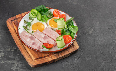 Breakfast plate with fried eggs, ham, fresh vegetables, and greens on a wooden board with copy space on dark background