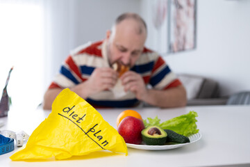 A man is eating junk food at a table. A yellow paper marked diet plan and a plate of fruit and...