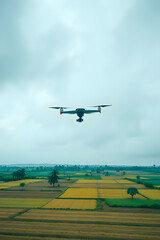 A high-angle view of a modern drone flying over a vibrant, checkerboard landscape of green and yellow agricultural fields. The sky is overcast and cloudy, creating a soft, even light.