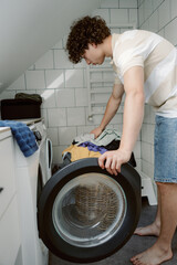 A young man in casual clothing is placing clothes into a washing machine in a well-lit laundry room. Sunlight streams through the window, highlighting his focused expression.