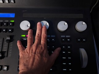 A person operating a professional lighting console panel with one hand, adjusting control knobs and buttons in a studio or live event setting.